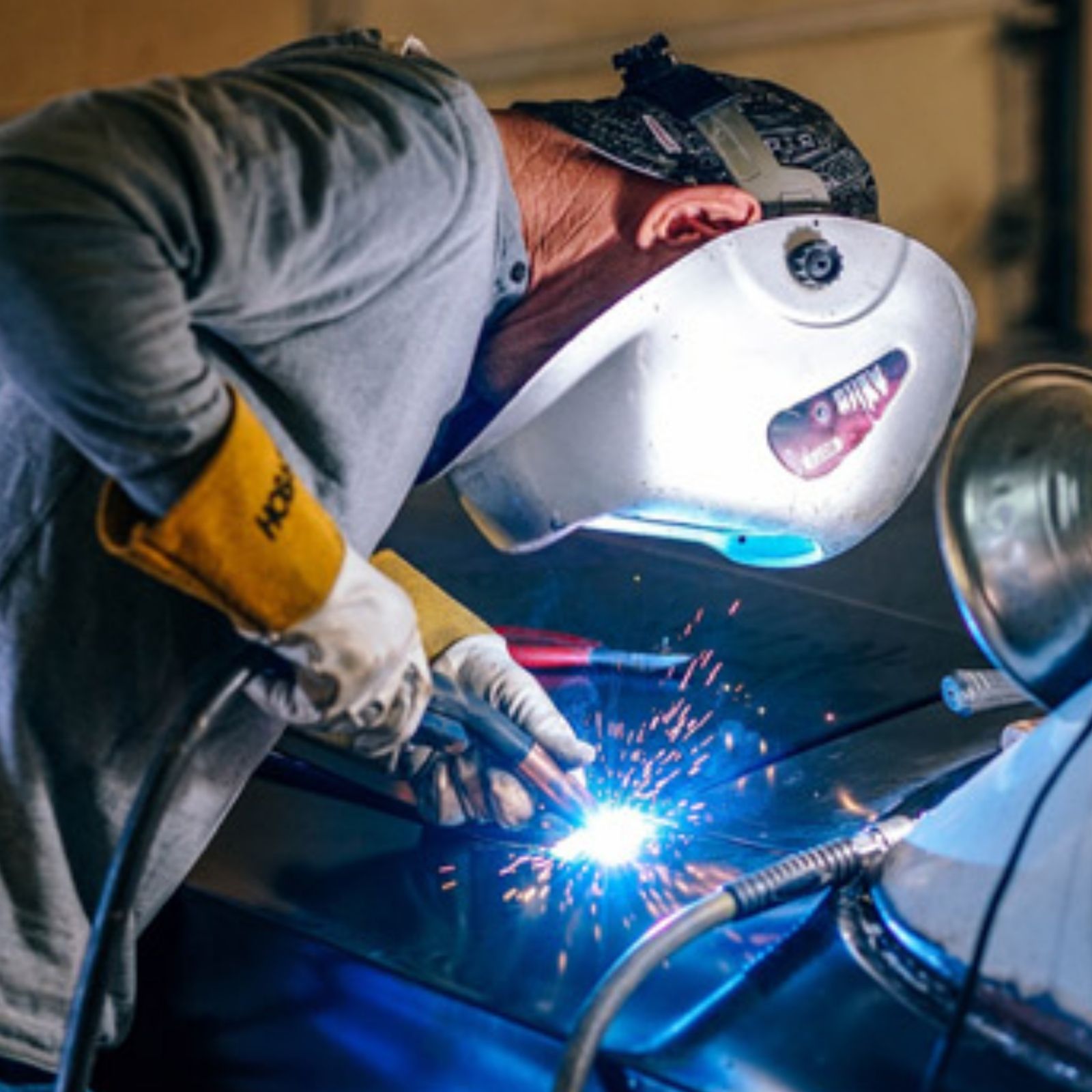 A person welding sheet metal repair to a parked car in a metal workshop.